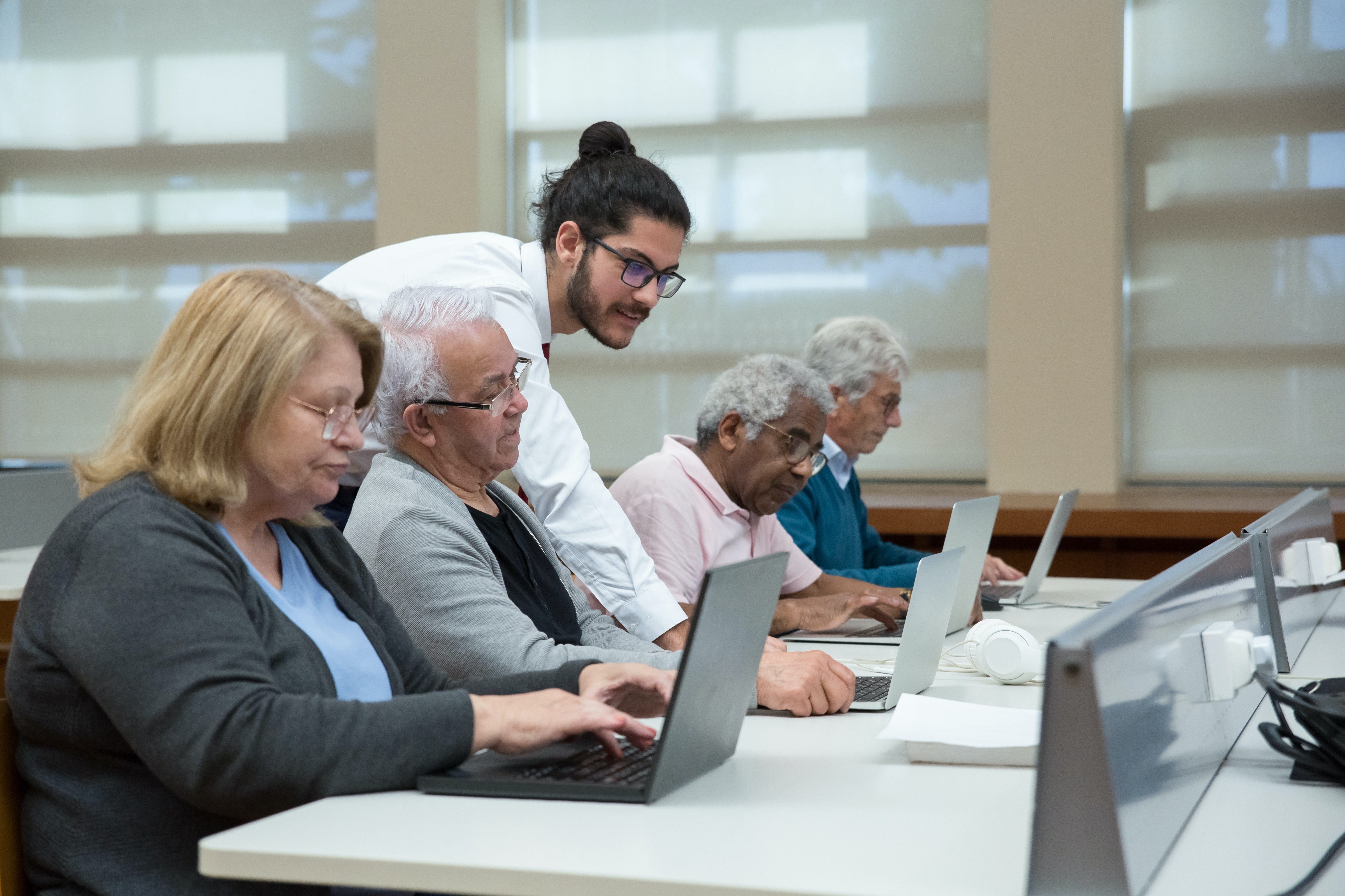 A man teaching a group of elderly people using laptops. The teacher is a younger man