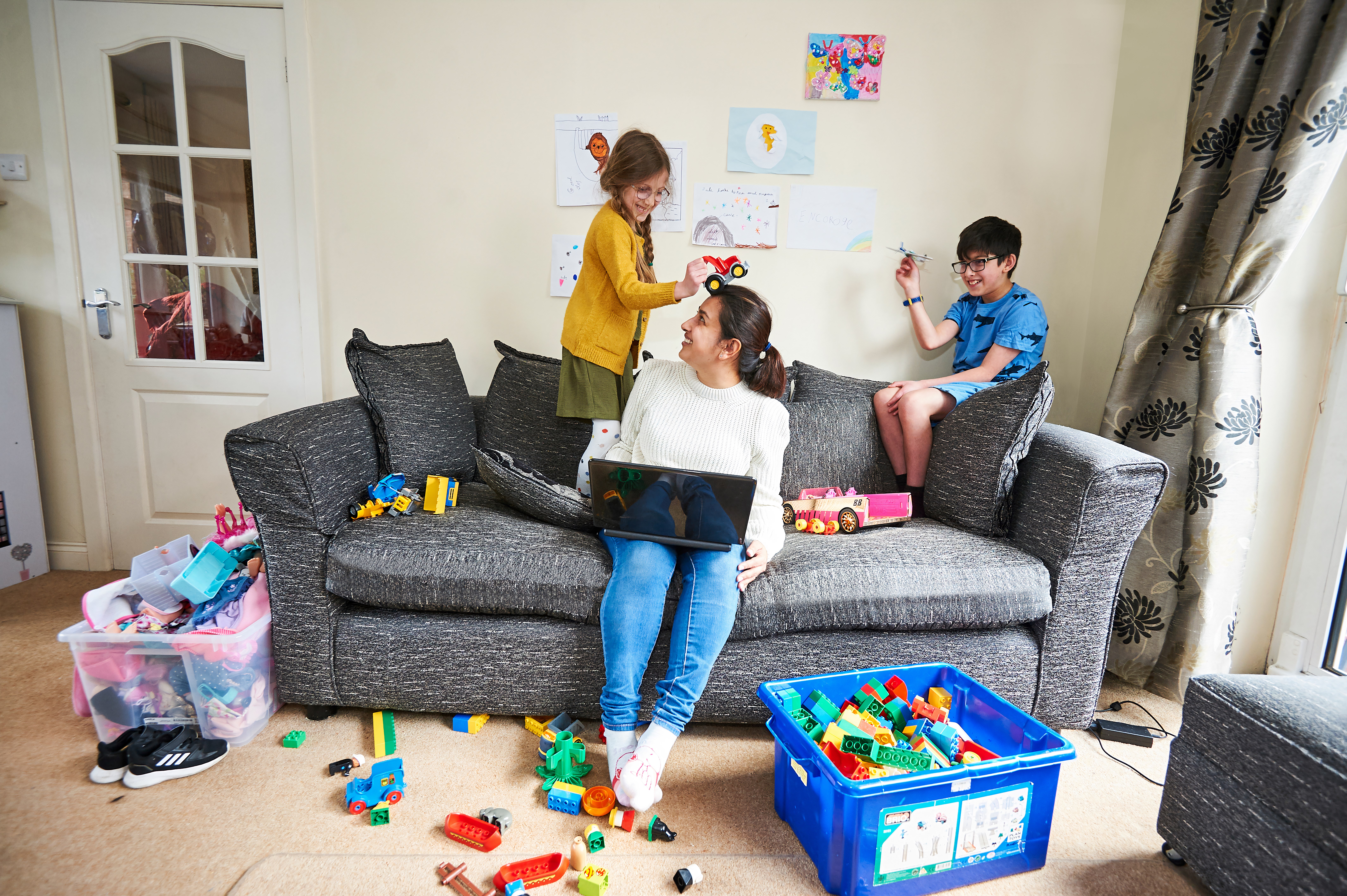 Woman sat on a sofa with her laptop and toys all around her, there are two children playing on the sofa. 