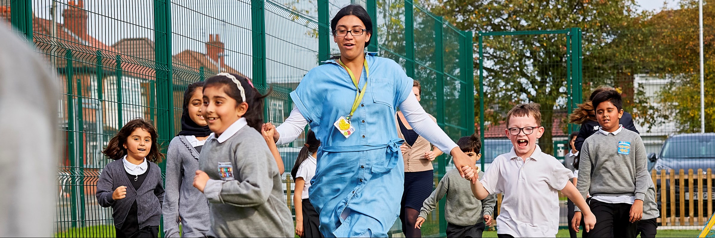 Children and adult running in a grassed school environment. Both the adult and children are smiling as they run.