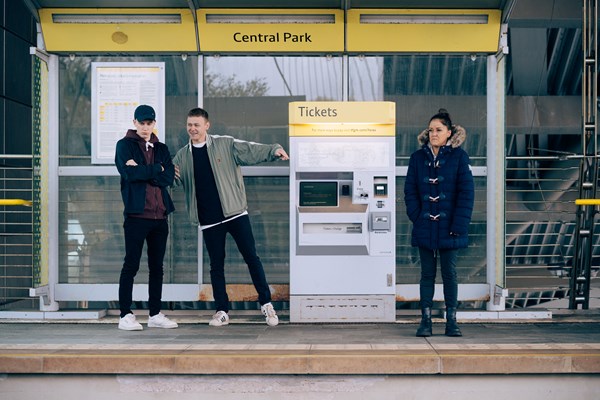 A young white man pointing at an older white woman at a Metrolink platform, whilst the young man's friend looks uncomfortable at his behaviour.