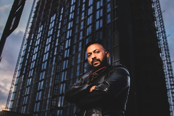 Photo of Sam Malik with the camera low down looking up at him with a high rise office block behind him, lit in a dramatic dark way. 