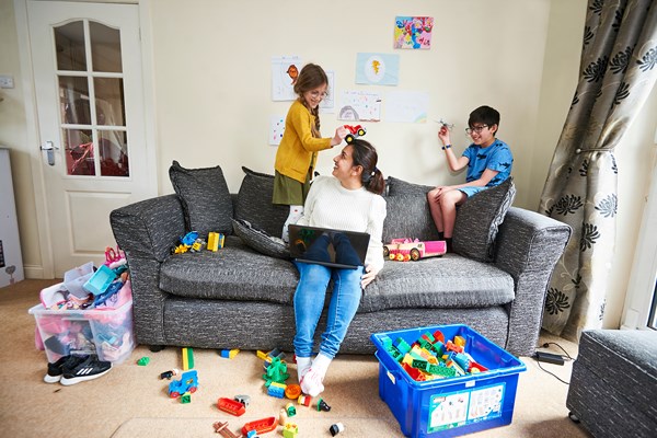 Woman sat on a sofa with her laptop and toys all around her, there are two children playing on the sofa. 