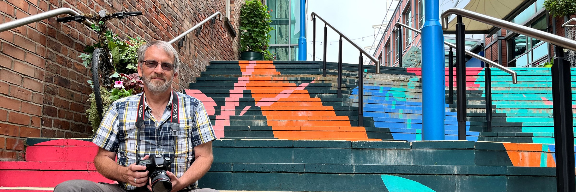 A person wearing a shirt holding a camera sitting on multicoloured steps.