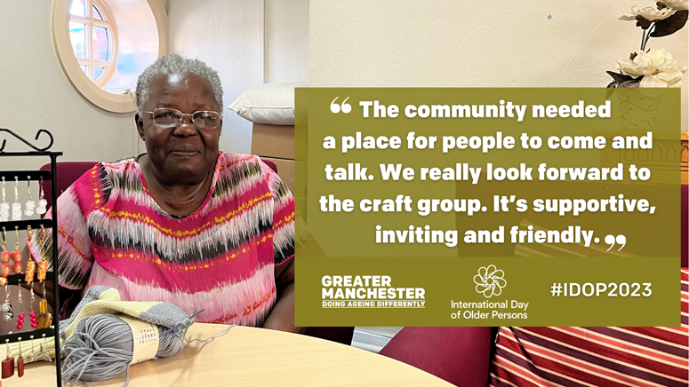 A person in glasses sitting behind a table with a jewellery rack and a ball of wool on it. Graphic text reads, ‘”The community needed a place to come and talk. We really look forward to coming. It’s supportive, inviting and friendly.”