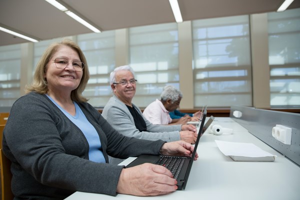 An older man and woman sit at a table with laptops. They are smiling at the camera