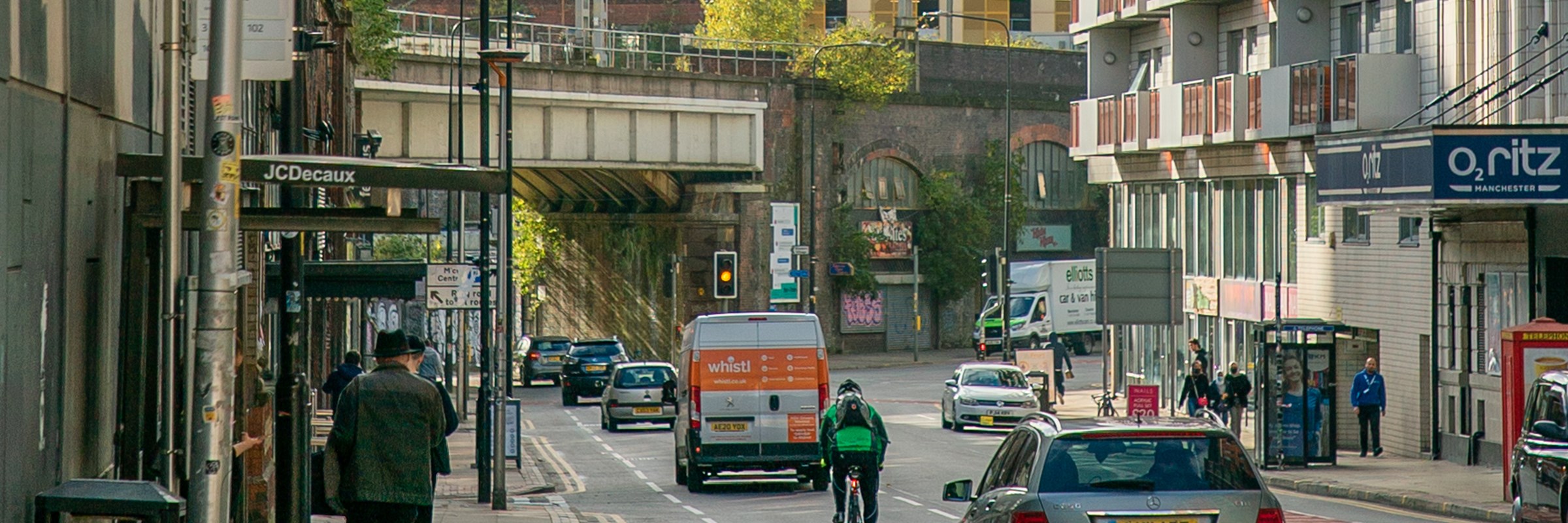 Busy road in Manchester city centre with pedestrians walking on pavements, cars and a cyclist use the road heading towards a bridge, buildings can be seen in the background.