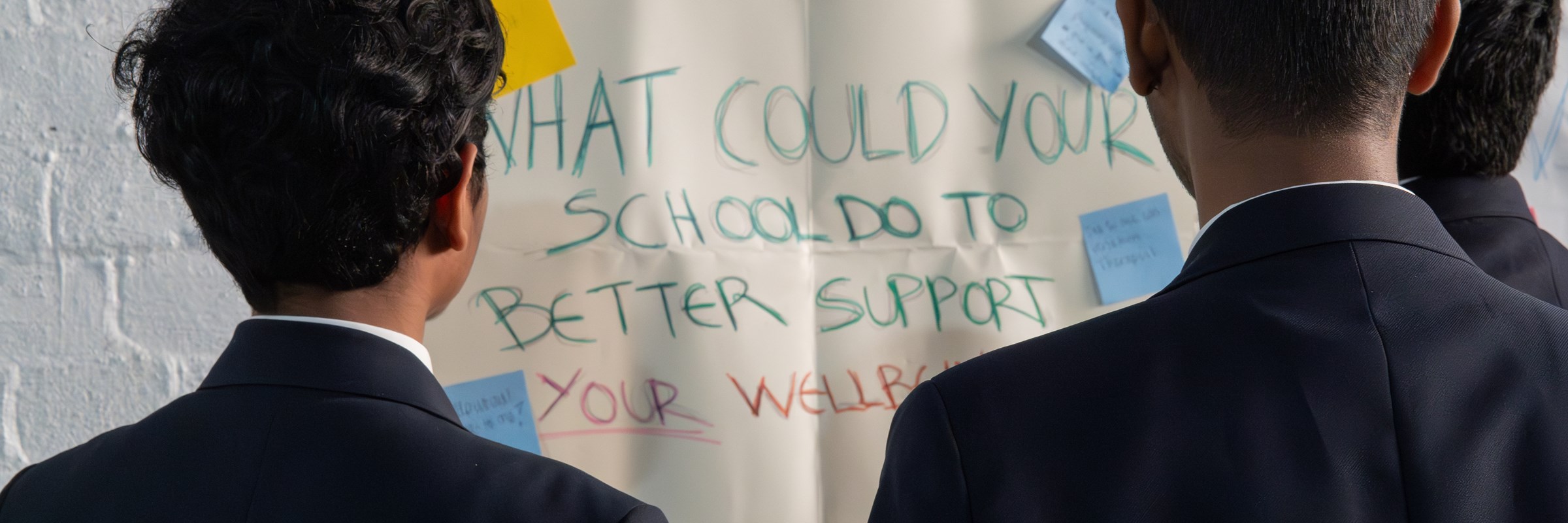 Three school children looking at a poster asking: 'What could your school do to better support you wellbeing?'