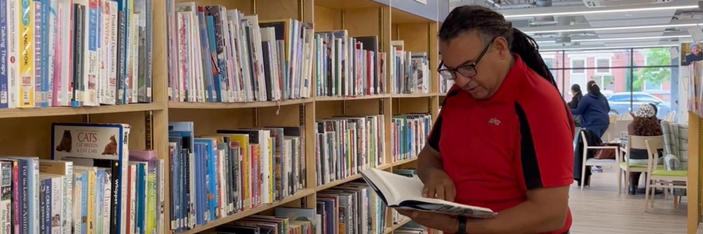 A person with long hair and glasses reading a book from a library.